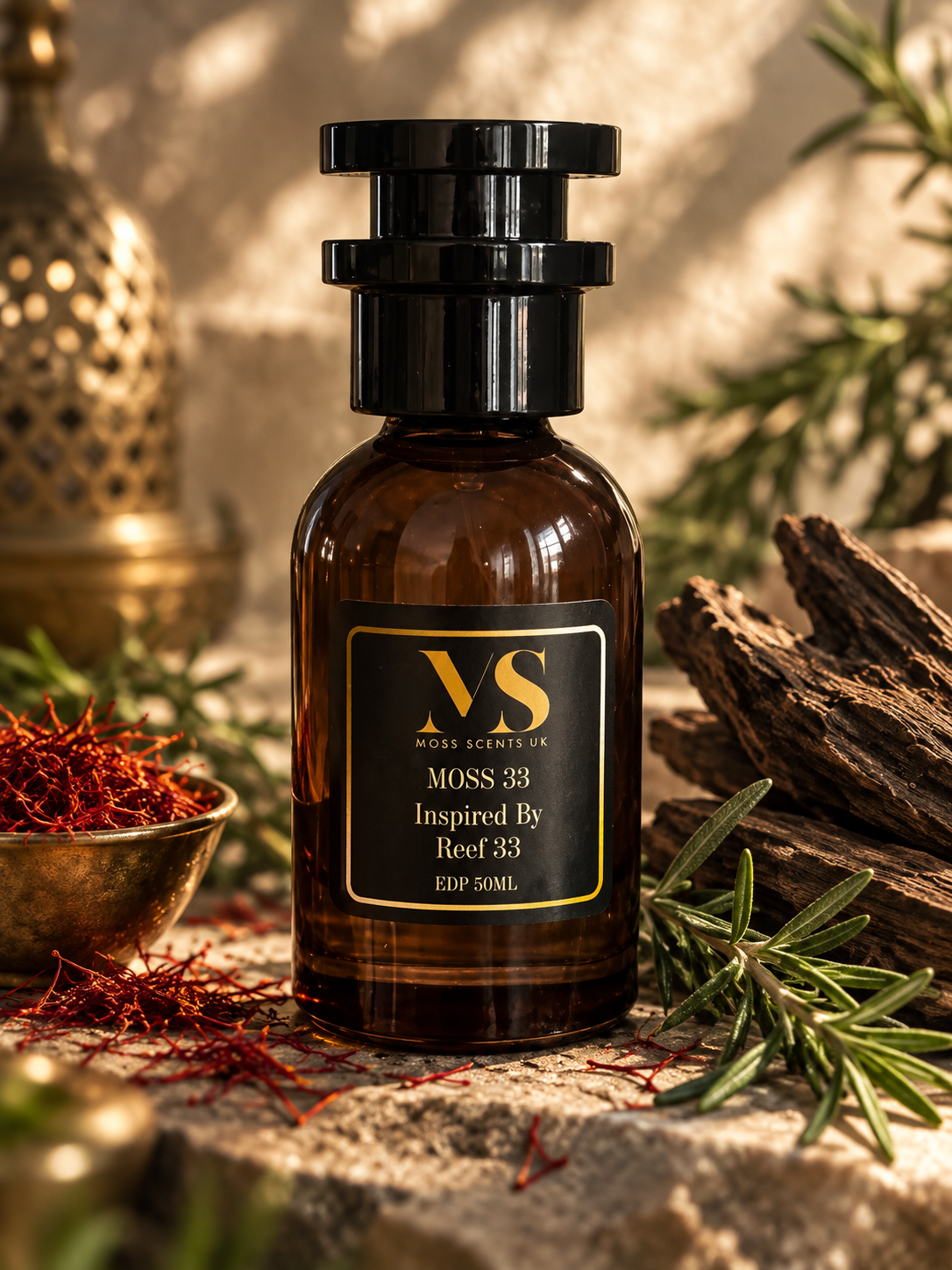 An amber glass bottle with a black cap and a black label reading ' MOSS SCENTS UK Inspired By Reef 33 ' in gold lettering, styled on a stone surface with saffron, rosemary, wood, and a brass vessel in the background.