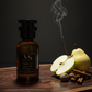 A bottle of inspired by red tobacco perfume with a lit incense stick, half-cut apple, pear, and cinnamon sticks on a wooden surface.
